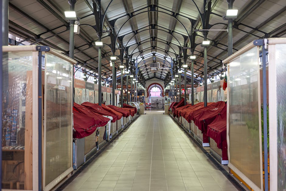 Interior view of a large market hall with a high, arched ceiling supported by black metal beams and a series of evenly spaced white ceiling lights. The floor is tiled with light brown and beige tiles, running down a central aisle. On both sides of the aisle, there are market stalls enclosed with transparent plastic panels and light wooden framing. Each stall features a flat surface covered with red fabric or tarps, some of which are draped over objects, possibly furniture or equipment. The stalls are arranged in rows, creating a symmetrical pathway leading to a bright arched window at the far end of the hall. The scene captures a moment of quiet, possibly during a moving or packing process, with furniture or items covered and prepared for transport, and the environment suggesting an active commercial or market setting aligned with house removals and furniture transport services by Brixton Removals.